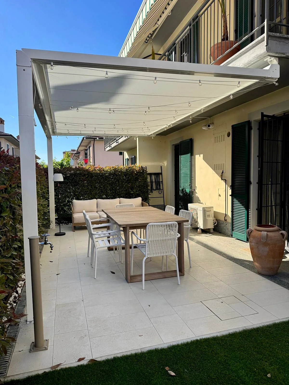 Pergola Tenso Block bianca con il telo steso sopra il tavolo da pranzo in un giardino, agganciata sotto il balcone